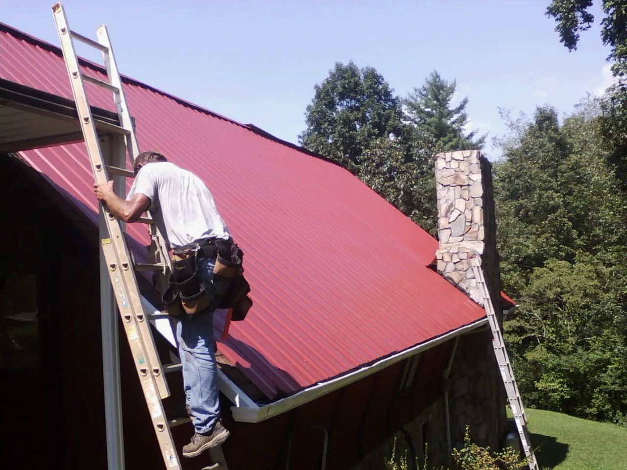 Roofer on a ladder beside a red metal roof and stone chimney — Soffit & Fascia Repair by Roofing Brothers in Drumore